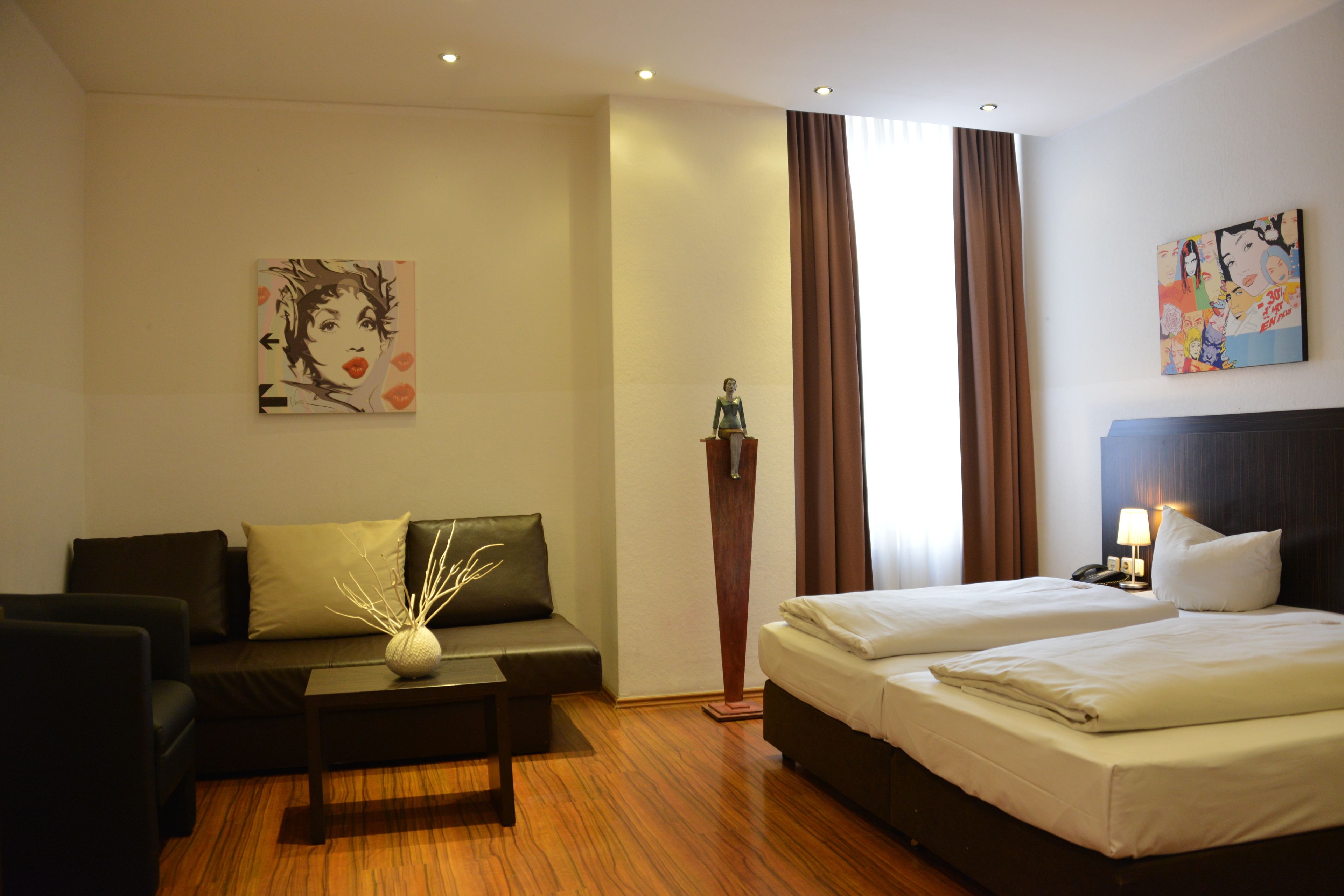 Habitación de hotel con sofá, mesa de centro, cama con dos almohadas, mesita de noche con lámpara y obras de arte en las paredes.