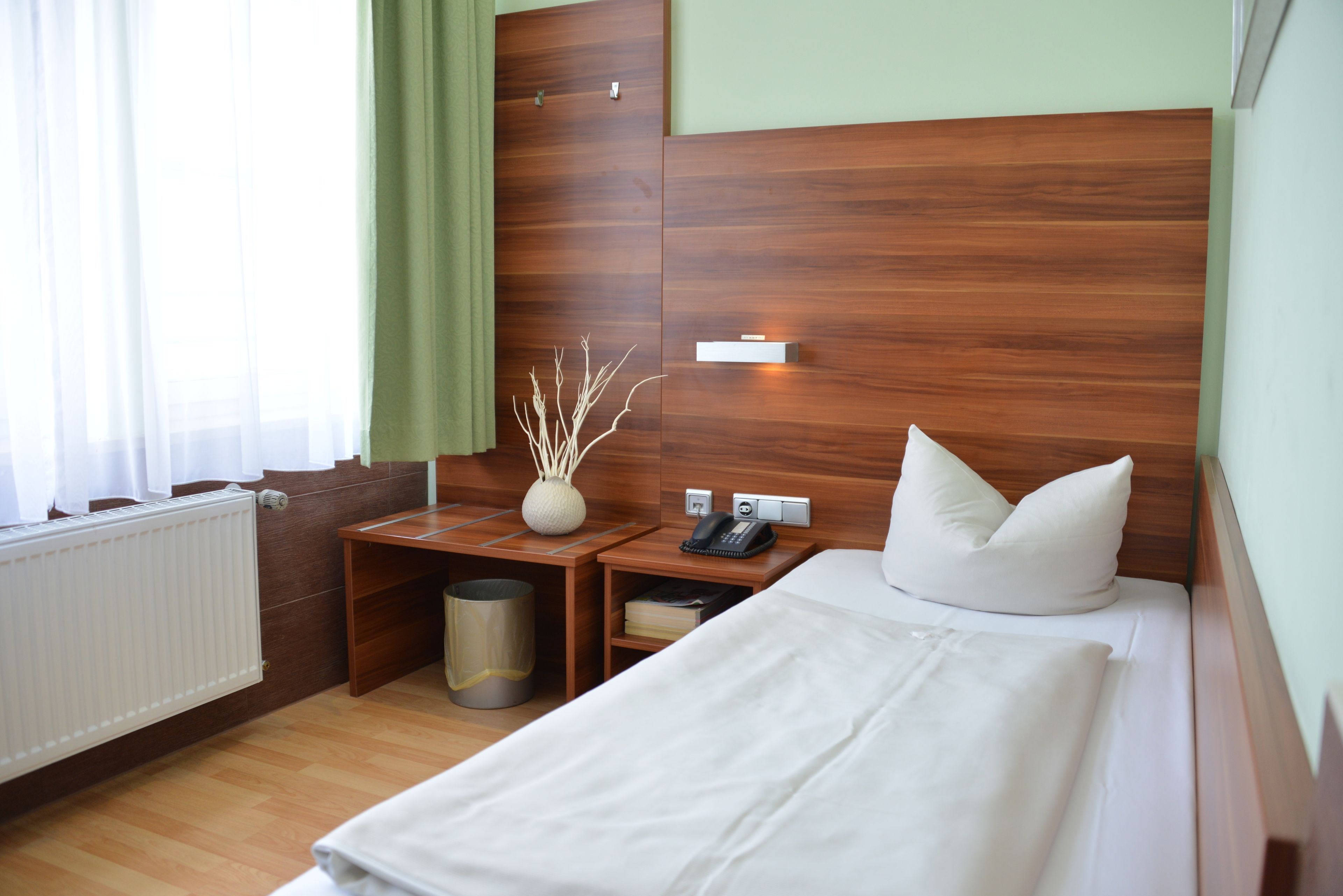 A hotel room with a wooden headboard, a bedside table with a lamp, a waste bin, and a bed with white bedding.
