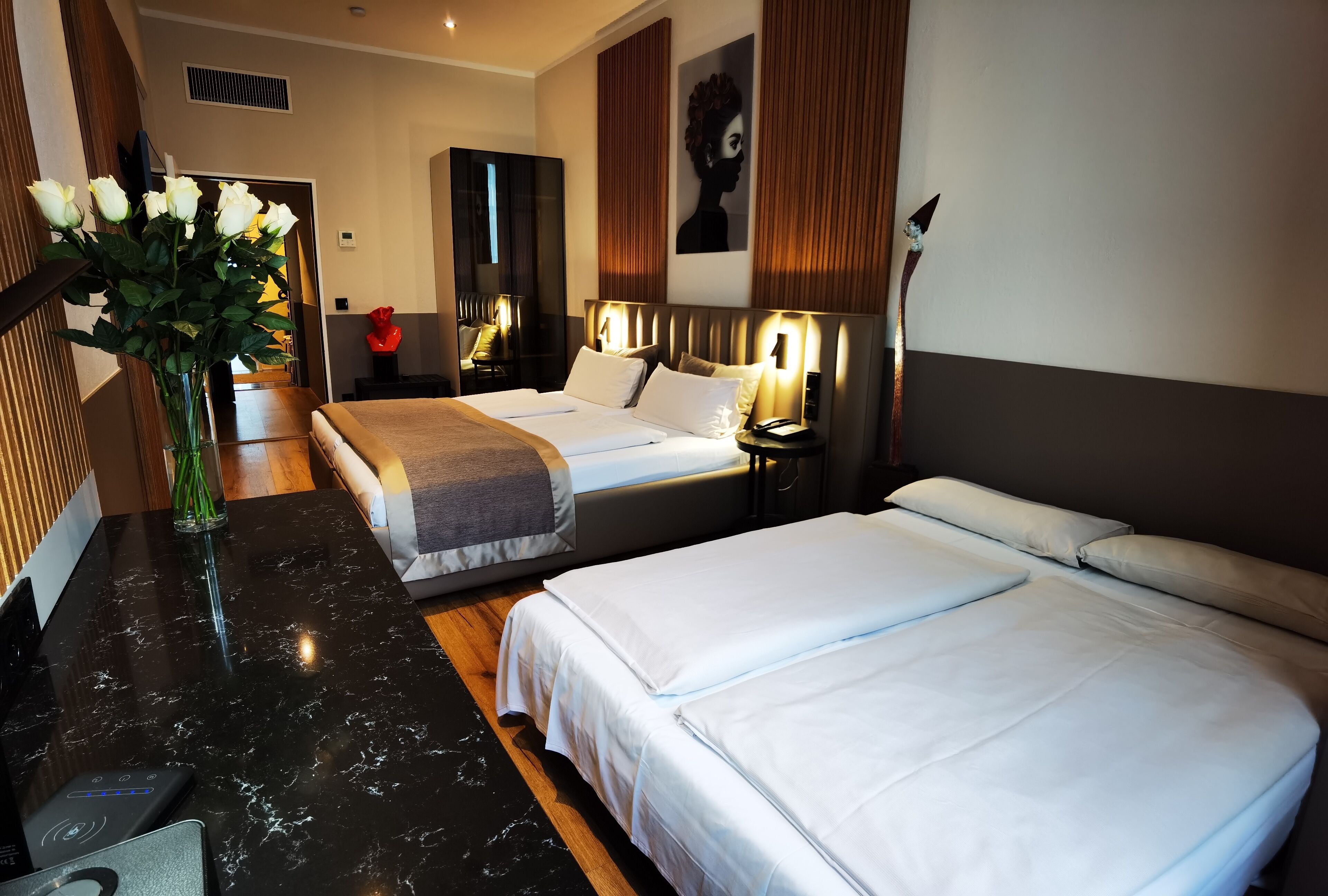 A hotel room with two beds, a marble-topped table, a vase with flowers, a mirror, and a red decorative item.