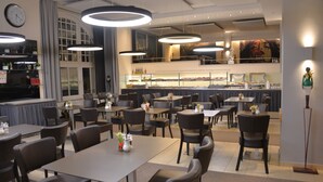 A restaurant with multiple tables and chairs, a counter with food display, and a clock on the wall. - Arthotel Munich (Munich)