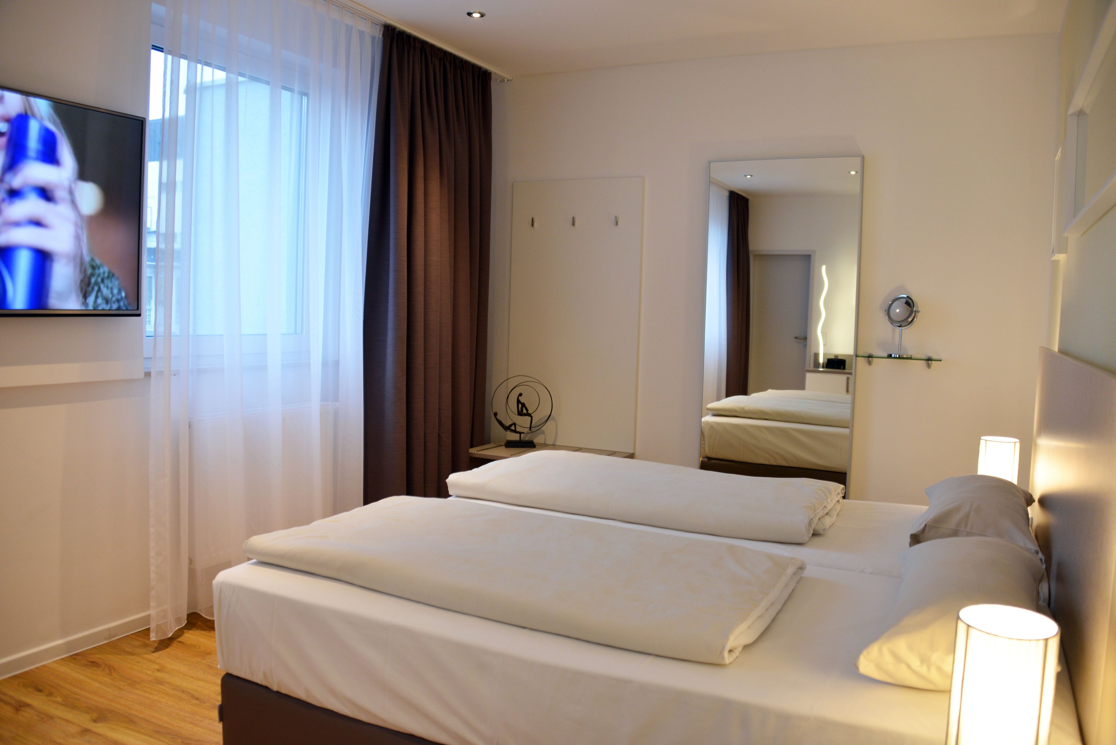 A hotel room with two beds, a TV, a mirror, and a bedside table with a lamp.