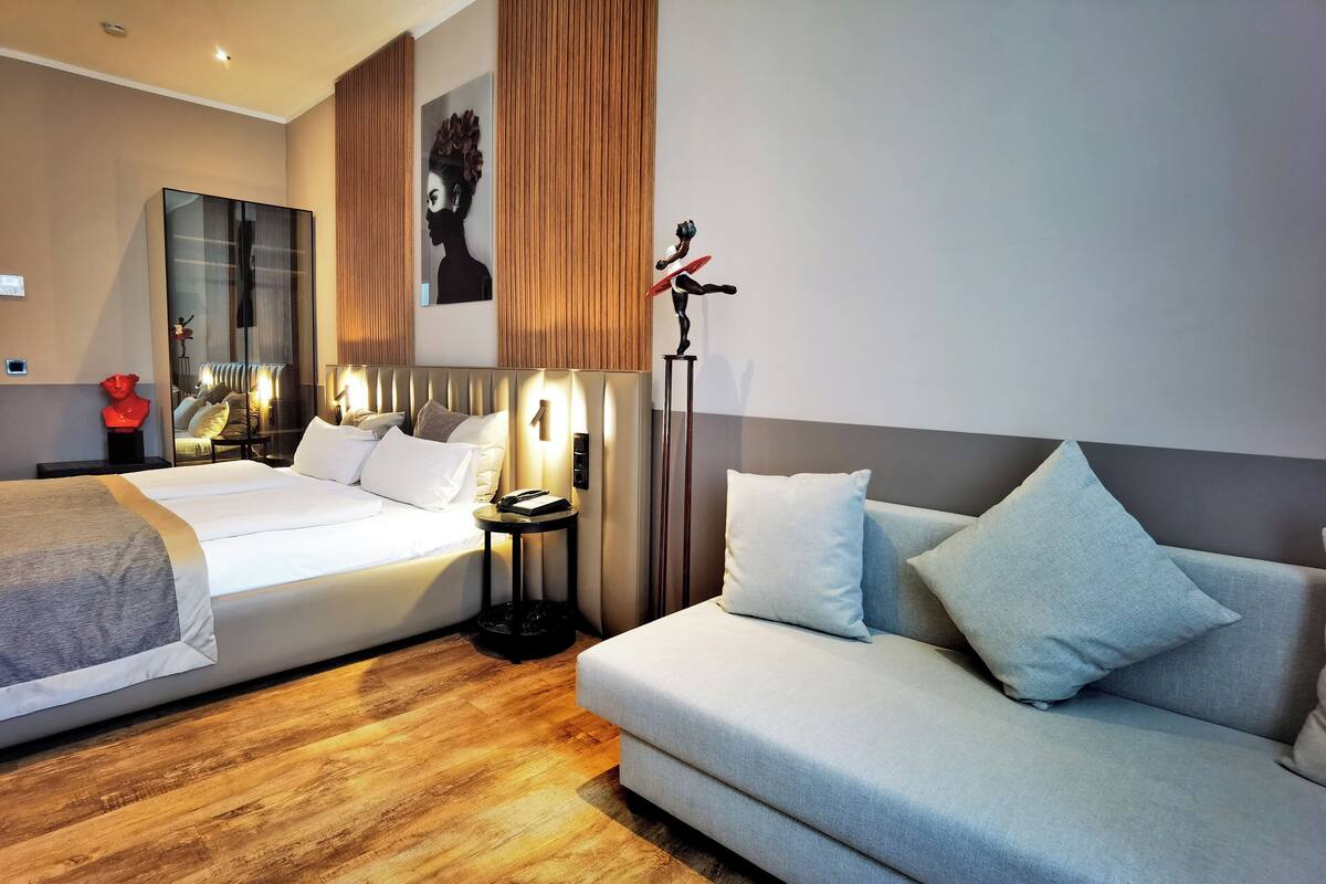 A modern hotel room with a large bed, a sofa, a nightstand, and a mirror.