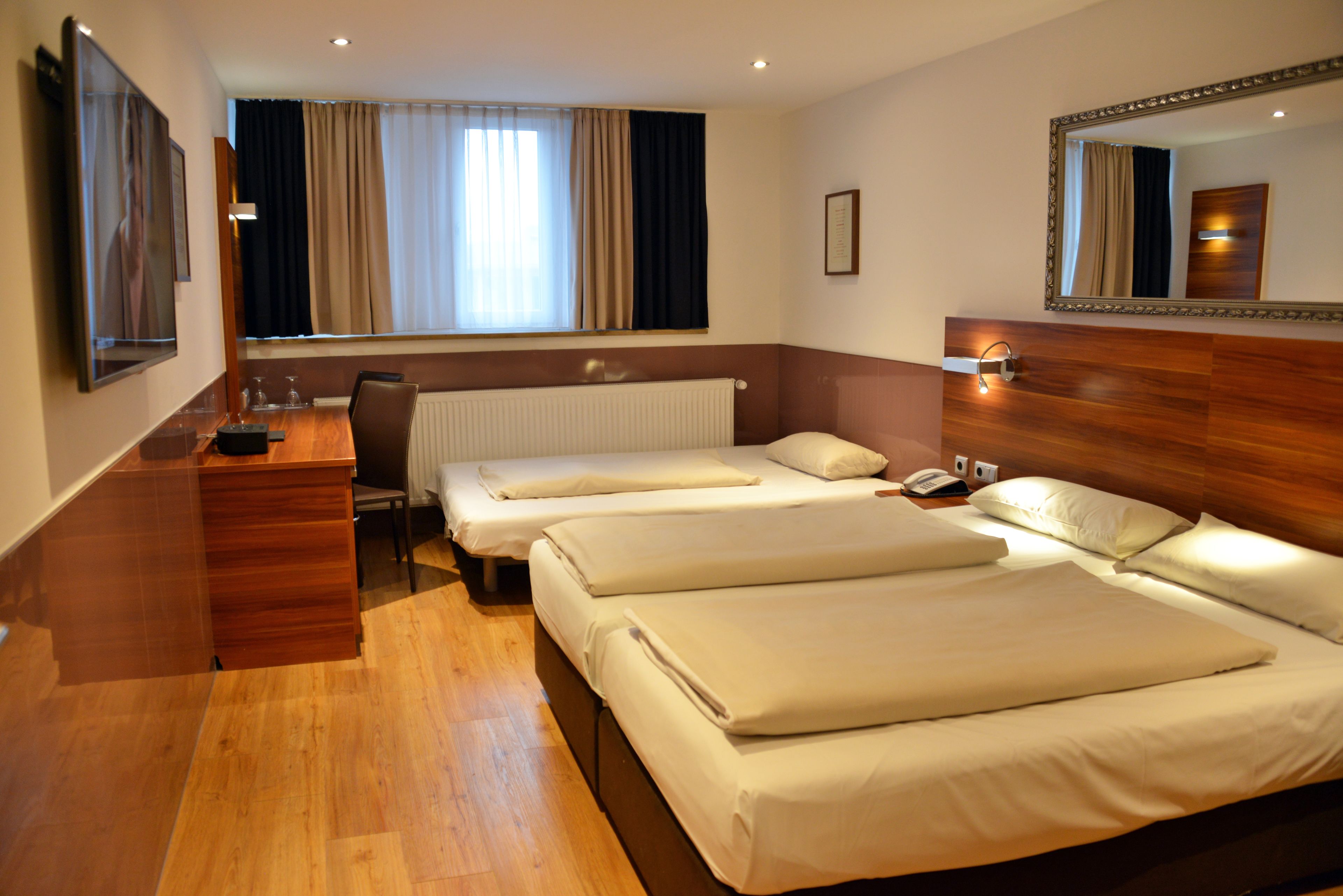 Ein Hotelzimmer mit zwei Einzelbetten, einem Schreibtisch mit Stuhl, einem großen Spiegel und einem Fenster mit Vorhängen.
