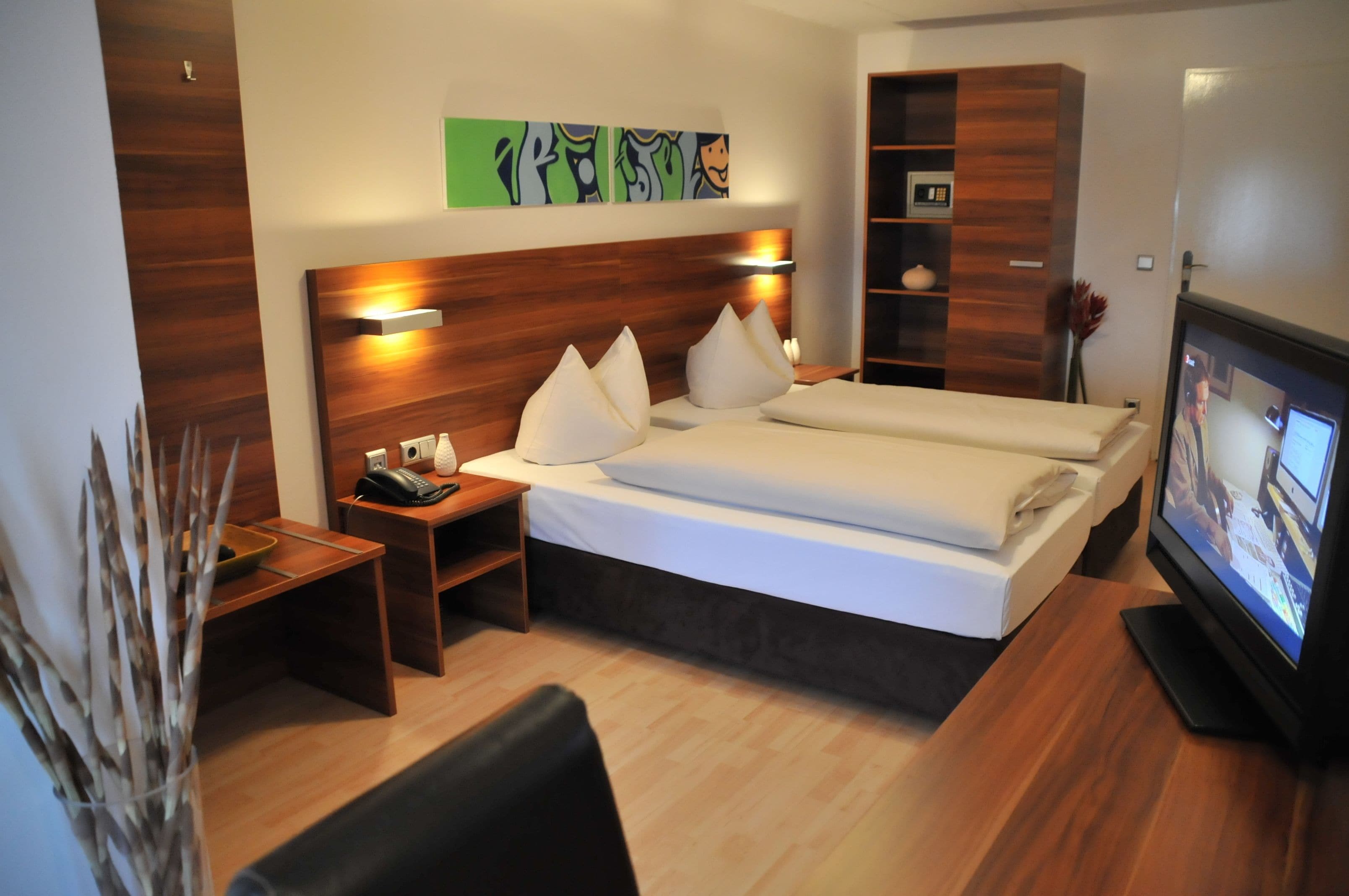 Une chambre d’hôtel avec une tête de lit en bois, un lit avec du linge de maison blanc, une table de chevet avec un téléphone, une télévision et une petite étagère avec des objets décoratifs.