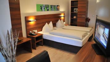 Une chambre d’hôtel avec une tête de lit en bois, un lit avec du linge de maison blanc, une table de chevet avec un téléphone, une télévision et une petite étagère avec des objets décoratifs.