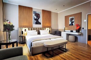 Una habitación de hotel moderna con una cama grande, mesitas de noche, un banco, una silla y obras de arte en las paredes.