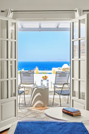 Eine geöffnete Tür, die zu einem Balkon mit Meerblick führt, mit zwei Stühlen, einem kleinen Tisch und einem blauen Teppich.
