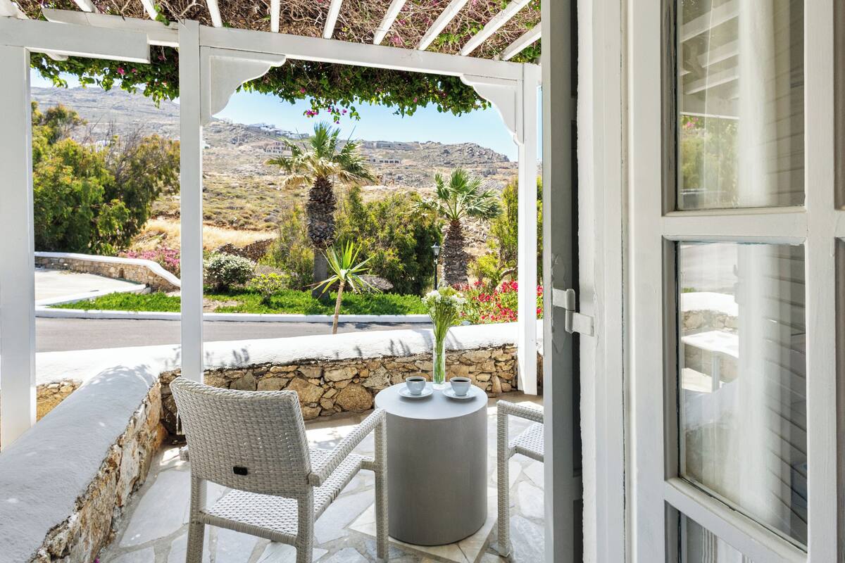 A covered outdoor seating area with a view of a mountainous landscape, stone walls, and a white pergola.