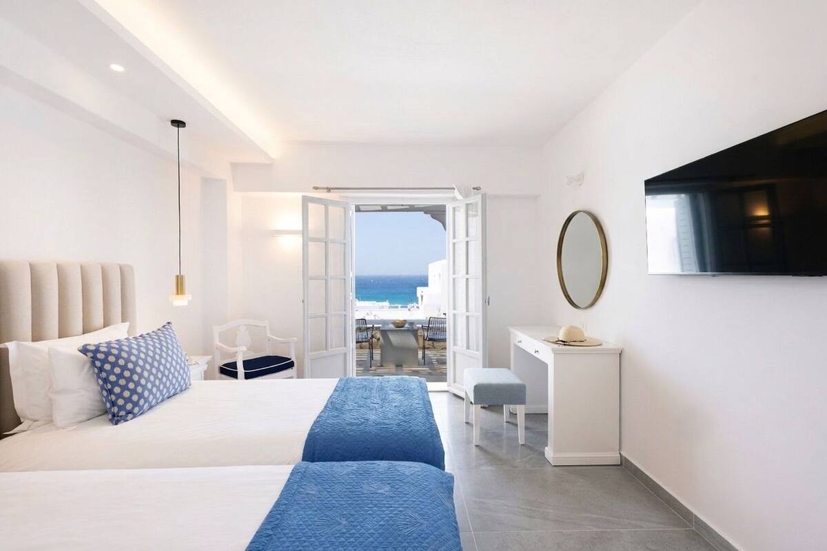 A hotel room with a bed, a desk, a chair, a TV, and a view of the sea.