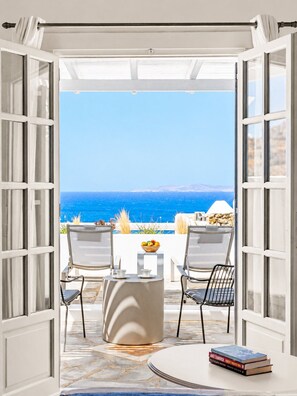 An open doorway leading to a balcony with two chairs, a small table, and a bowl of fruit. - San Marco Hotel (Mykonos)