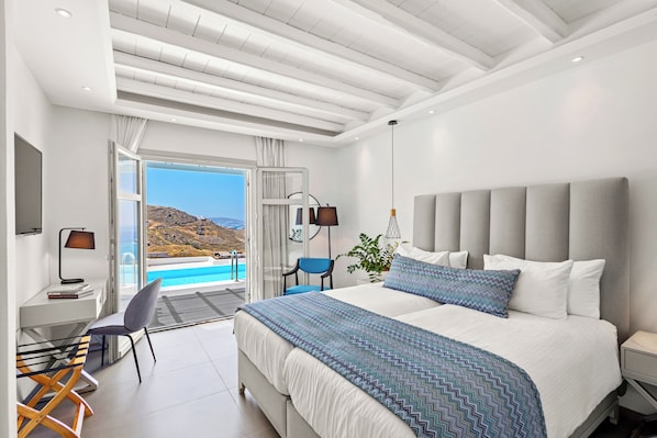 Ett modernt hotellrum med en stor säng, ett skrivbord och en stol. Genom den öppna dörren har man utsikt över en pool och berg.