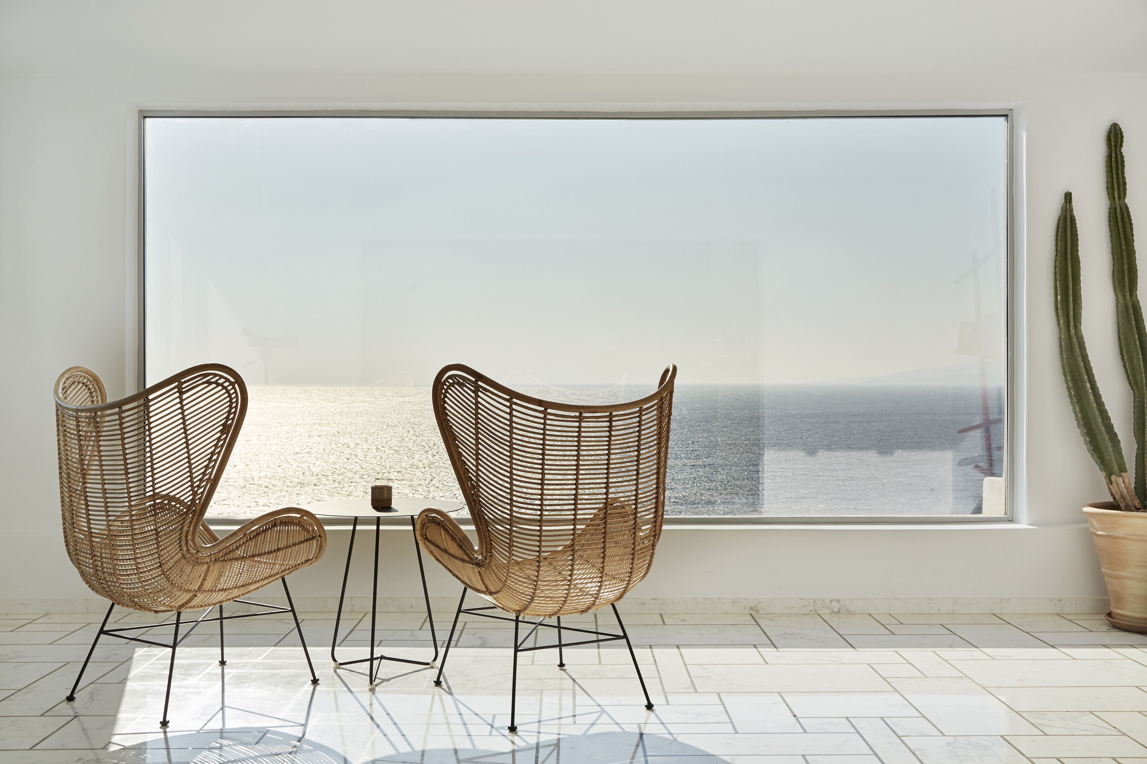 A room with two wicker chairs, a small table, and a large window overlooking a beach.