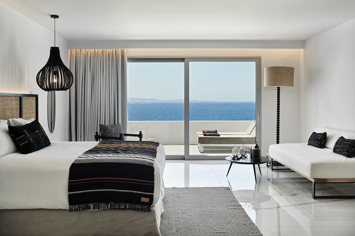 A modern hotel room with a large bed, a sofa, a balcony with a view of the sea, and a floor lamp.