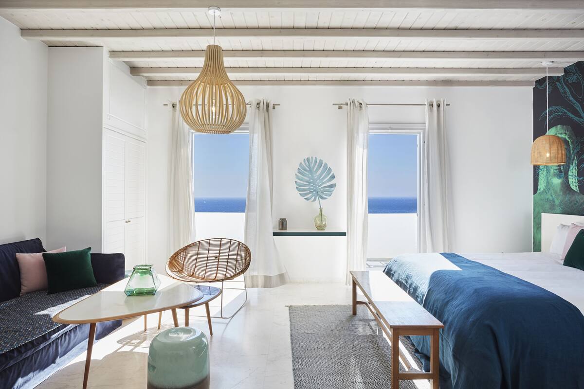 A bedroom with a bed, a sofa, a table, and a chair. There are two windows with curtains, a wall art piece, and a view of the sea.