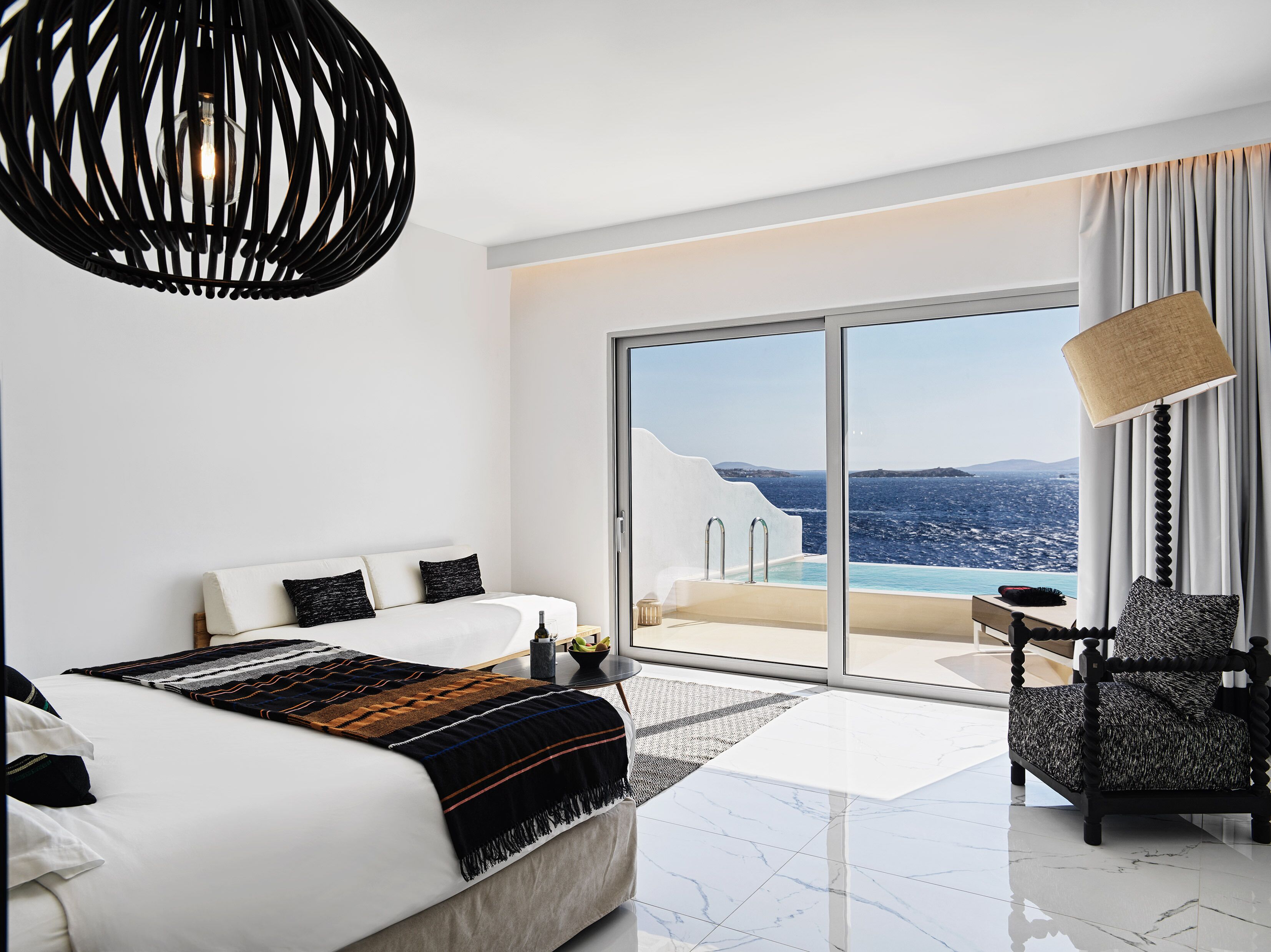 A modern hotel room with a large bed, a chair, a small table, and a view of the sea through a glass door.