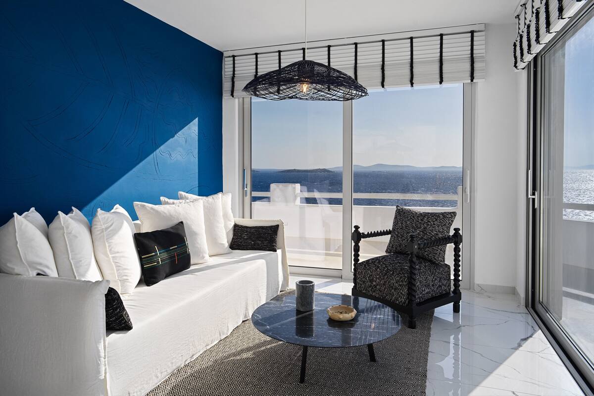 A modern living room with a white sofa, black and white patterned armchair, and a round table with a bowl. A large window offers a view of the sea and distant hills.