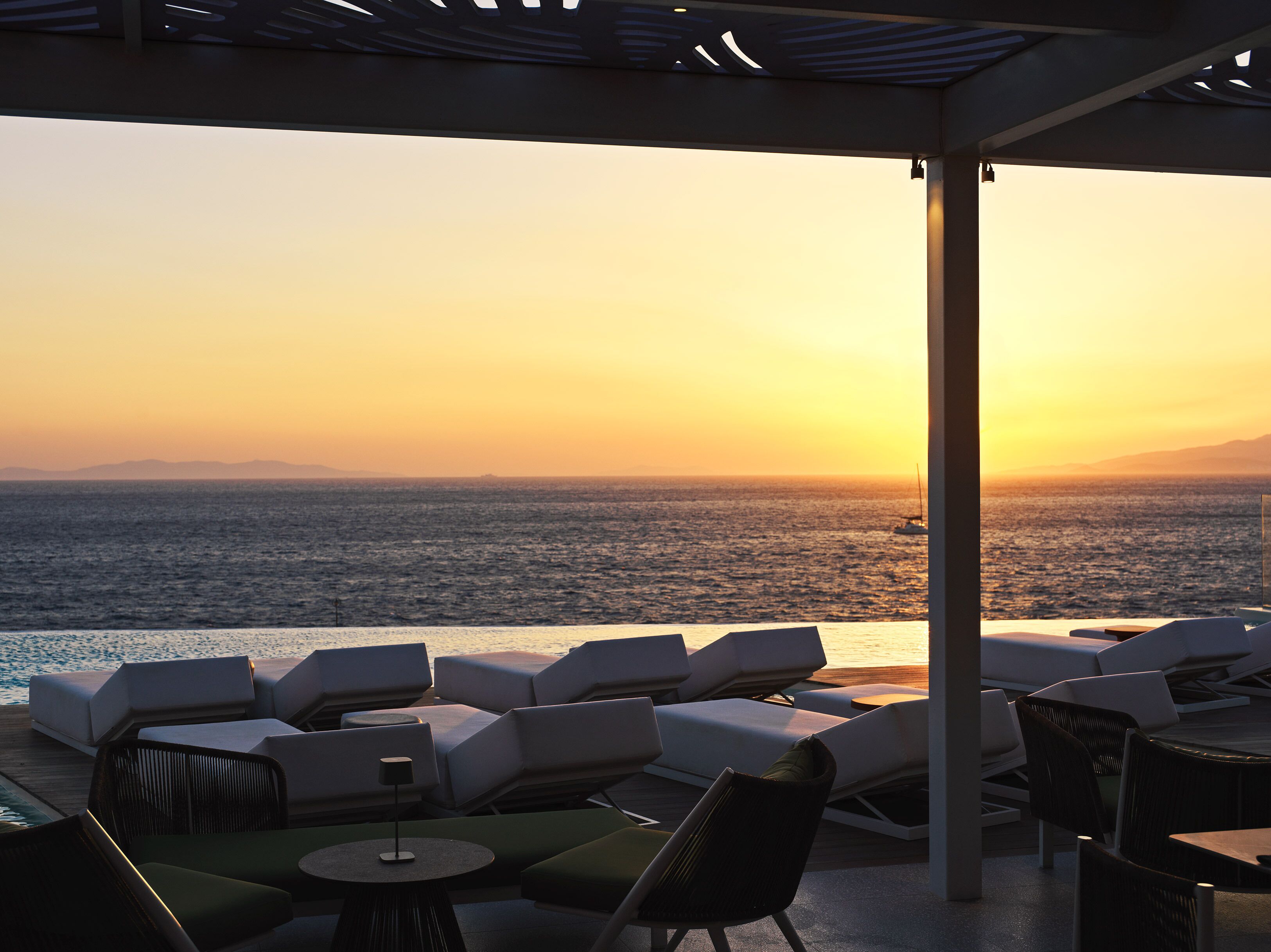 A sunlit outdoor seating area with lounge chairs and tables, overlooking a body of water at sunset.