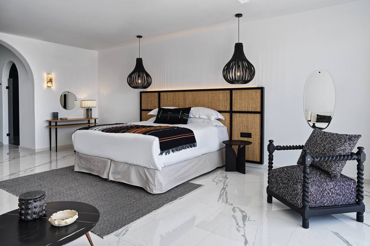 A modern bedroom with a large bed, two pendant lights, a round mirror, a small table, and a patterned armchair.