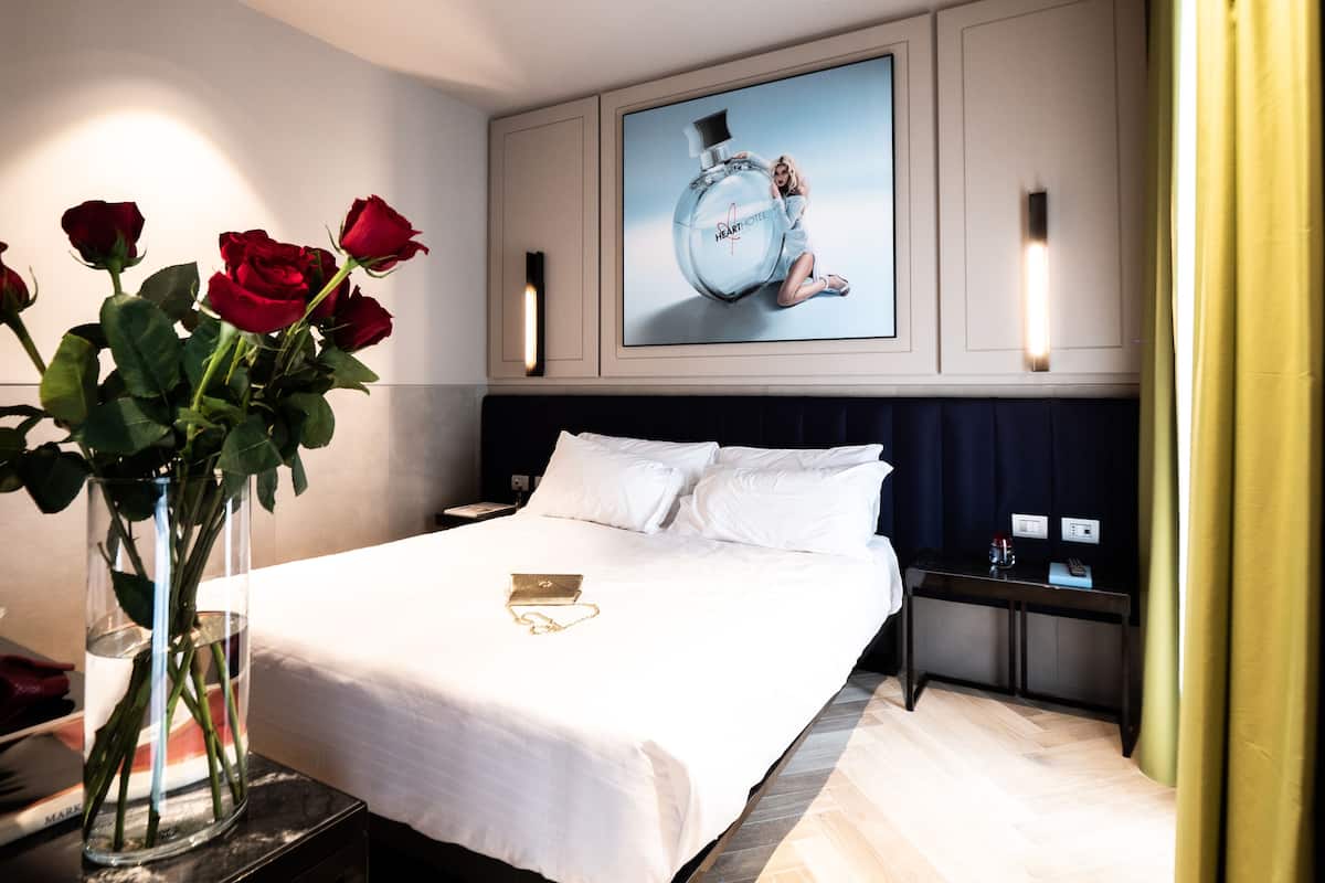 A hotel room with a large bed, white bedding, a vase with red roses, a nightstand, a lamp, and a poster on the wall.