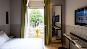 Une chambre d’hôtel avec un lit, un bureau, une télévision et un balcon donnant sur des arbres et un immeuble.