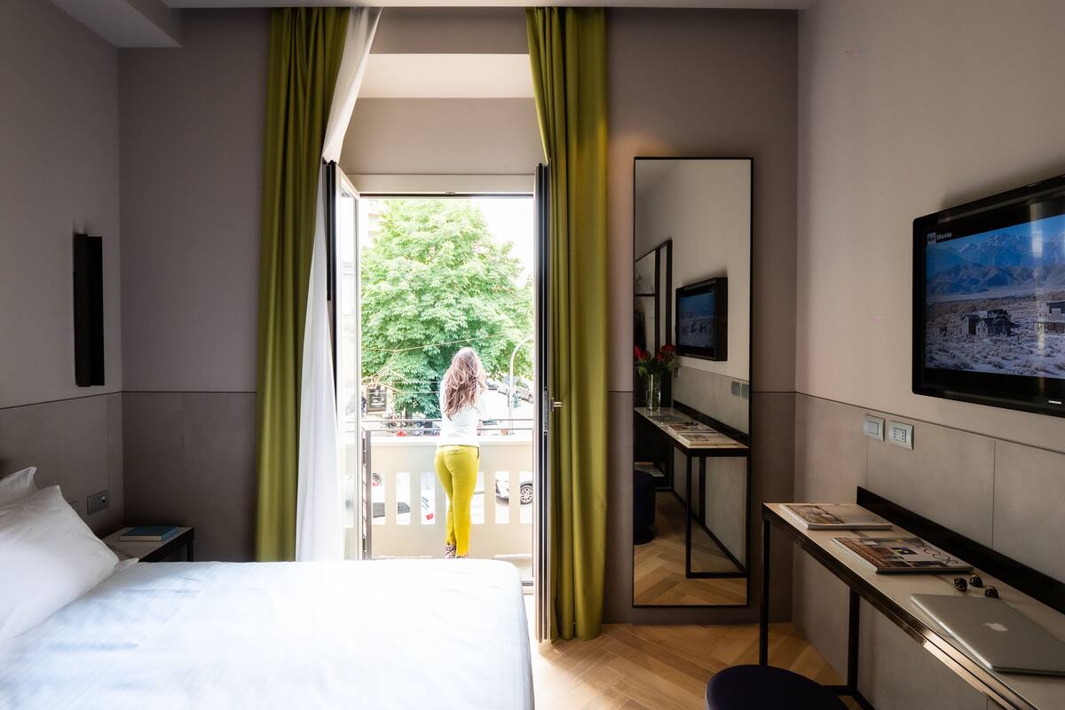 A hotel room with a bed, a desk, a TV, and a balcony with a view of trees and a building.