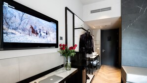 Une chambre d’hôtel moderne équipée d’un téléviseur à écran plat fixé au mur, d’un ordinateur portable posé sur un bureau, d’un vase avec des fleurs rouges et d’un porte-manteau sur lequel est accroché un vêtement noir.