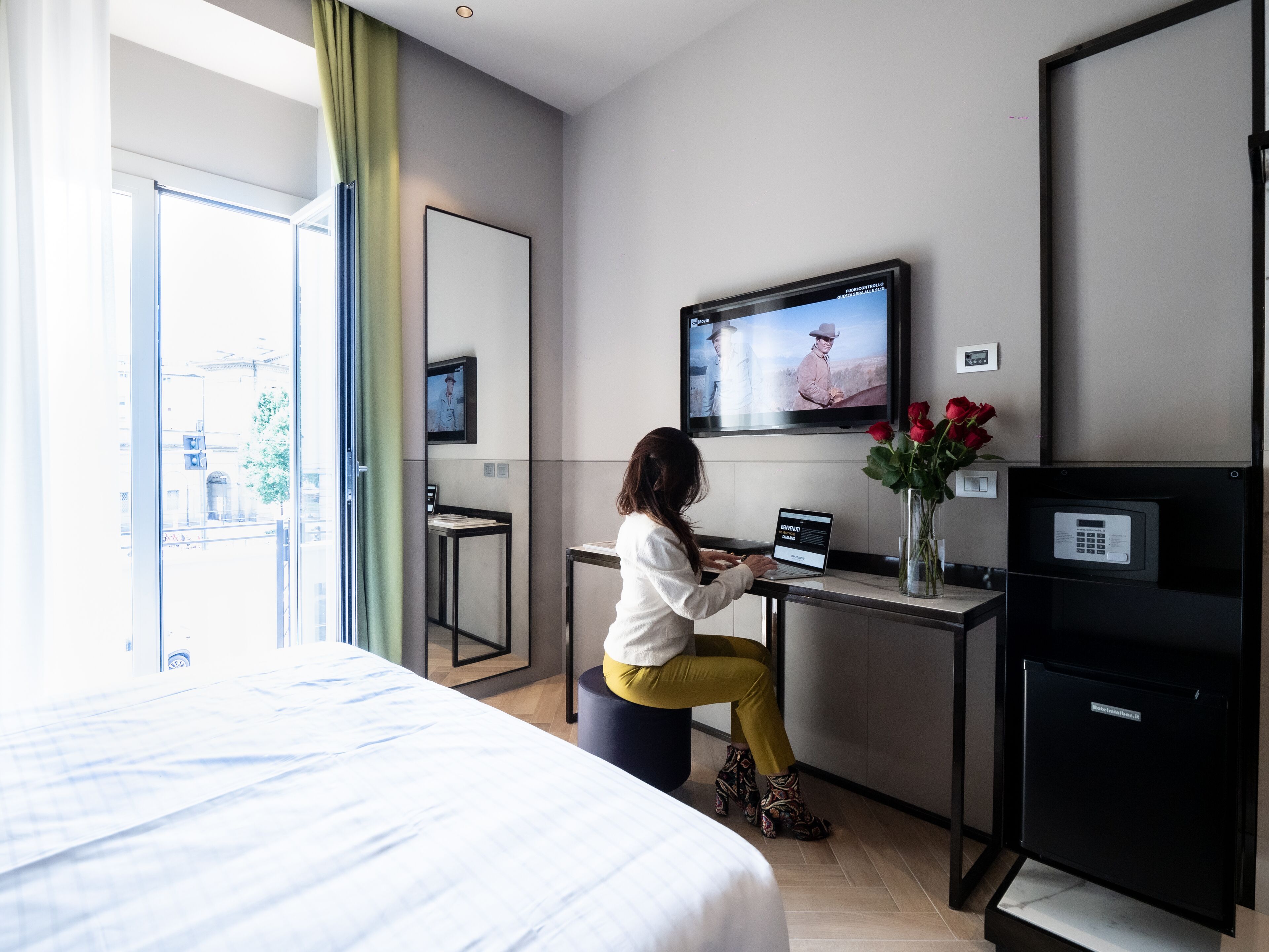 A hotel room with a bed, a desk with a laptop, a TV, and a vase with flowers.