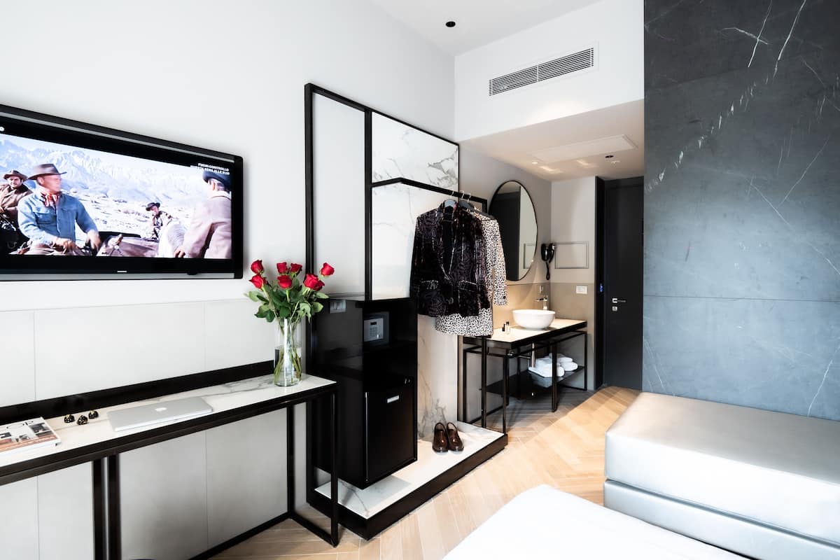 A modern hotel room with a flat-screen TV mounted on the wall, a black refrigerator, a vanity with a mirror, a sink, and a bed.