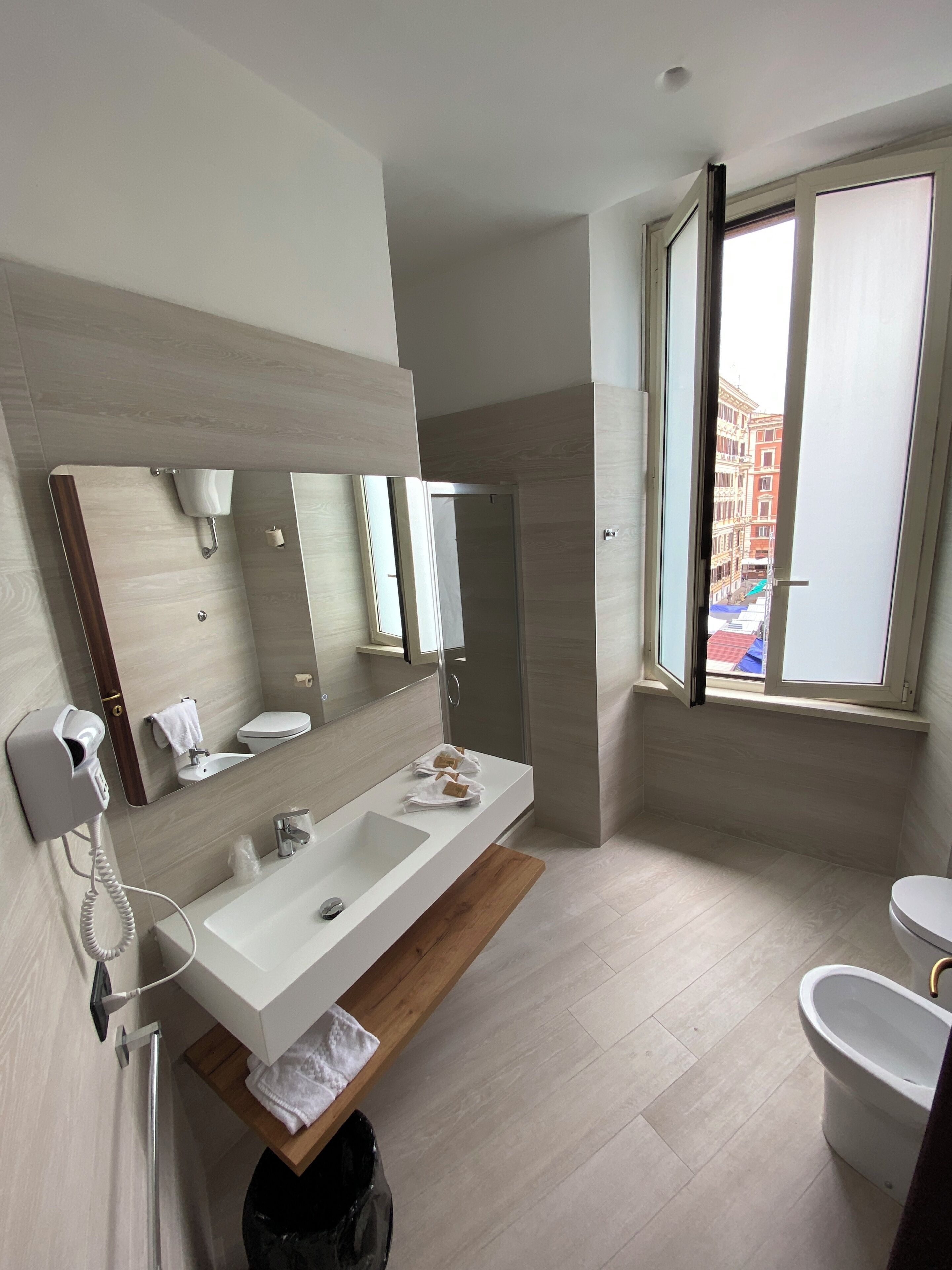 A modern bathroom with a large mirror, a white sink, a toilet, and a window with a view of buildings.