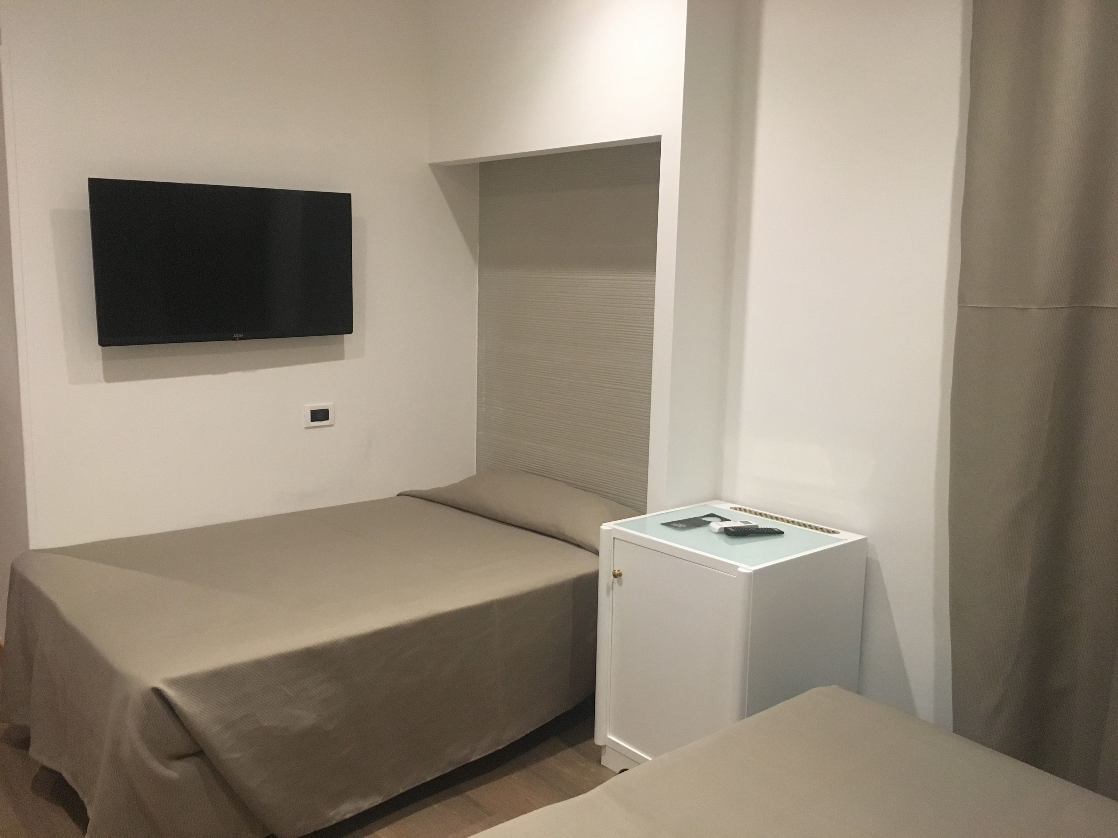 A small, compact room with two beds, a TV mounted on the wall, a small cabinet, and a window with blinds.