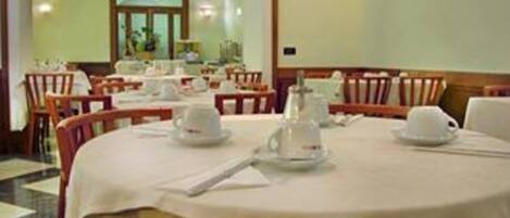 Comedor con mesas redondas dispuestas para cuatro comensales, cada una con mantel blanco, platos y tazas.