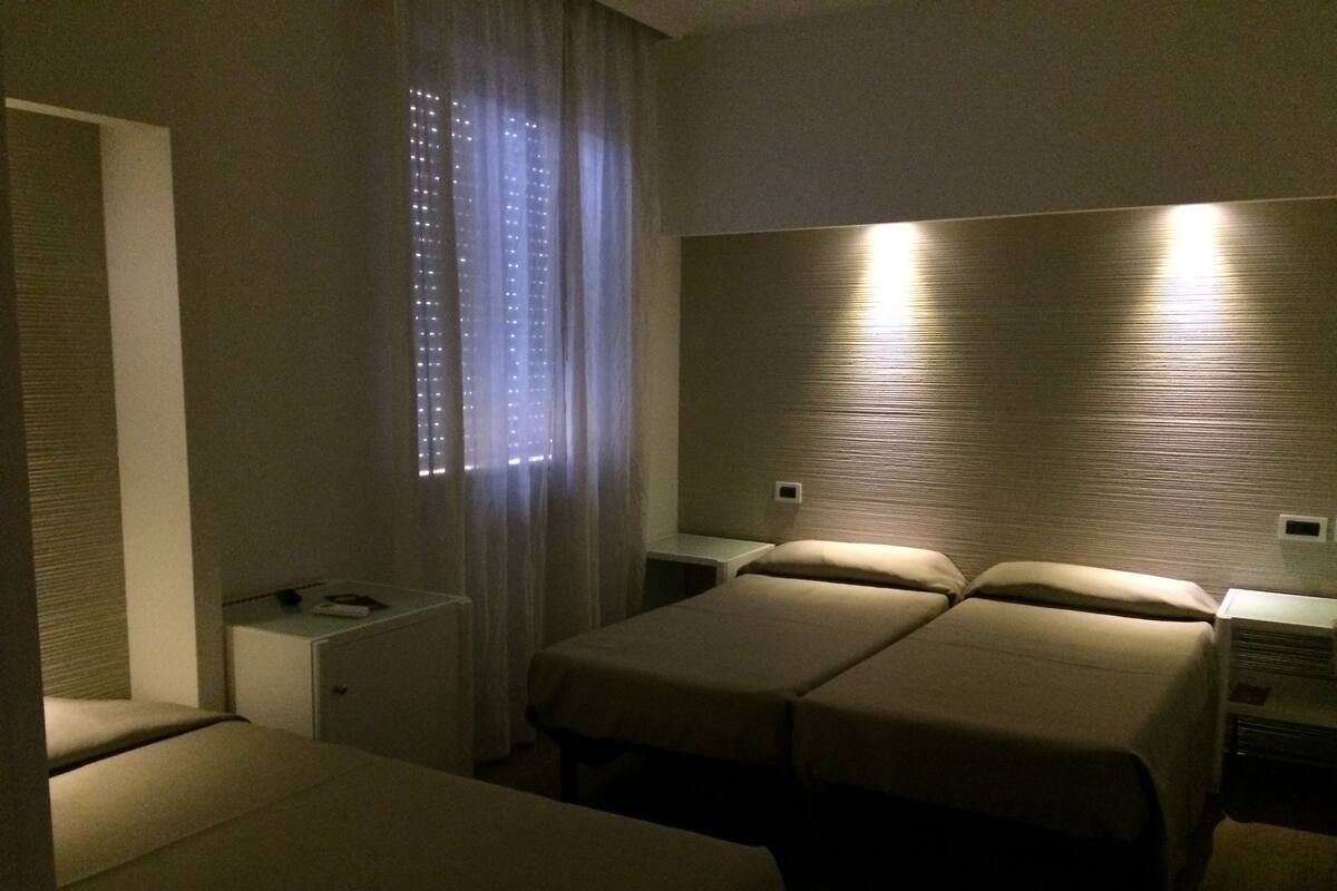 Quarto de hotel com duas camas de solteiro, uma mesinha de cabeceira e uma janela com cortinas.