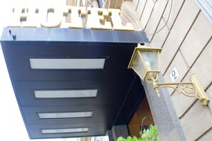 Un letrero de hotel con la palabra 'HOTEL' en letras grandes, una entrada moderna con fachada negra y un elemento decorativo de iluminación.