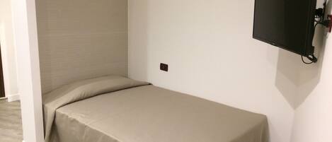 A small, single-bed room with a plain headboard, a mounted television, and a power outlet on the wall.