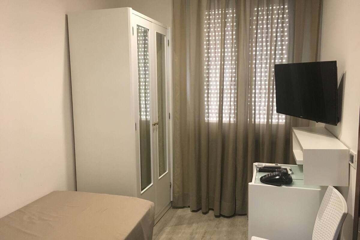 Quarto de hotel com cama, escrivaninha, televisão de tela plana, guarda-roupa e janela com cortinas.