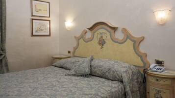 A bedroom with a bed, two framed pictures on the wall, a nightstand with a phone, and wall sconces.