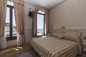 A bedroom with a large bed, a view of the sea through a window, and a television mounted on the wall.