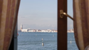 A view through a window with curtains, revealing a body of water with several buoys and a distant cityscape with buildings and a tower. - Hotel Riviera (Venice)
