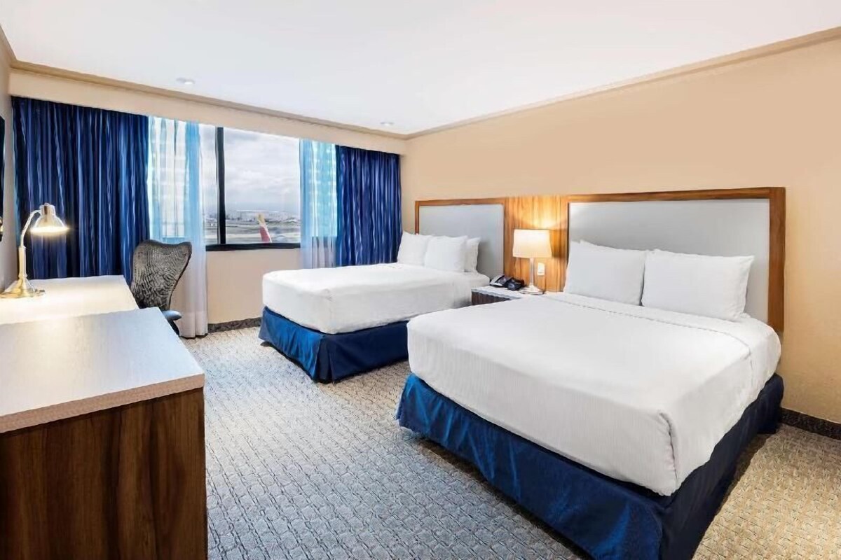 Une chambre d’hôtel avec deux lits, un bureau, une chaise et une grande fenêtre avec des rideaux.