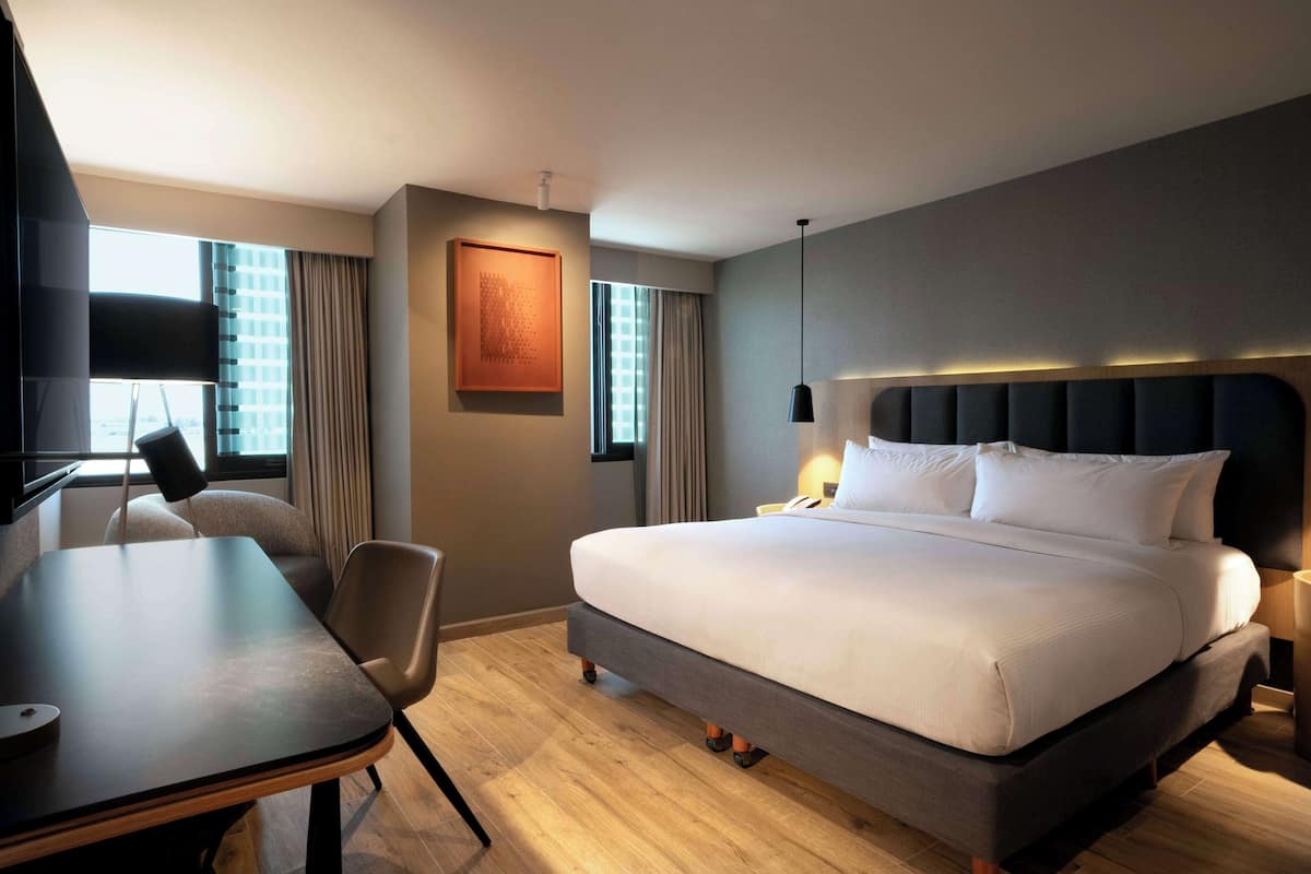 Habitación de hotel con una cama grande, un escritorio y una silla. Hay una ventana con cortinas, un cuadro en la pared y una lámpara en la mesita de noche.