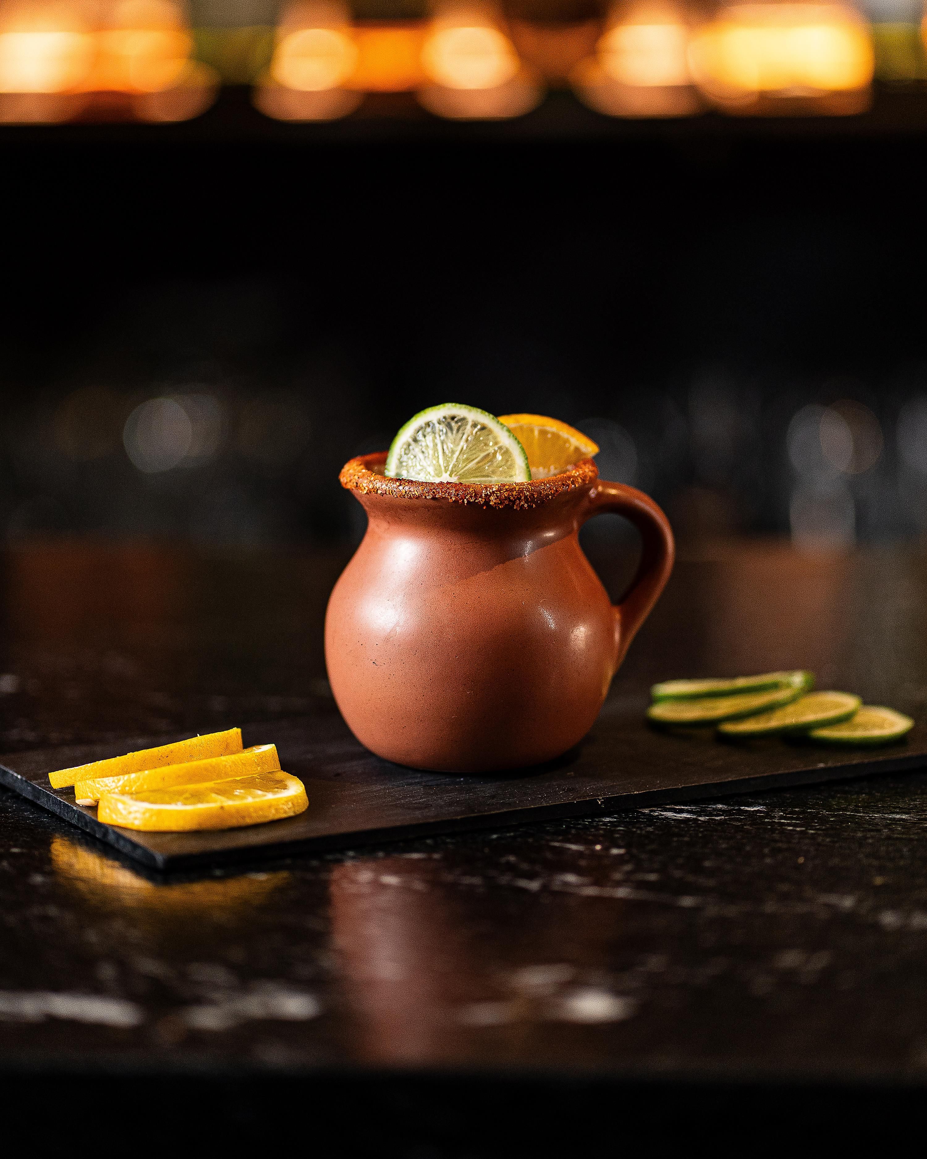 A terracotta pitcher with a lime slice on top, surrounded by orange slices on a dark surface.