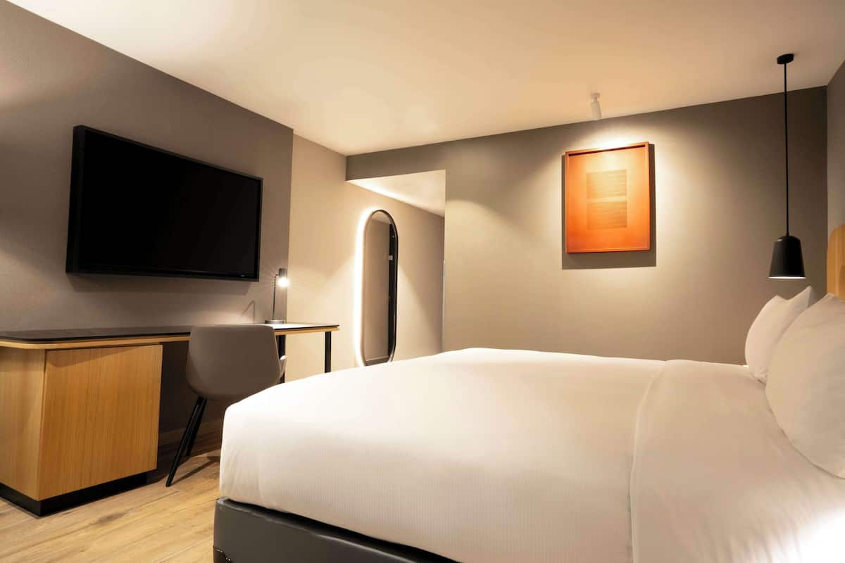 Una habitación de hotel moderna con una cama grande, un escritorio, un televisor de pantalla plana en la pared y una lámpara colgante.
