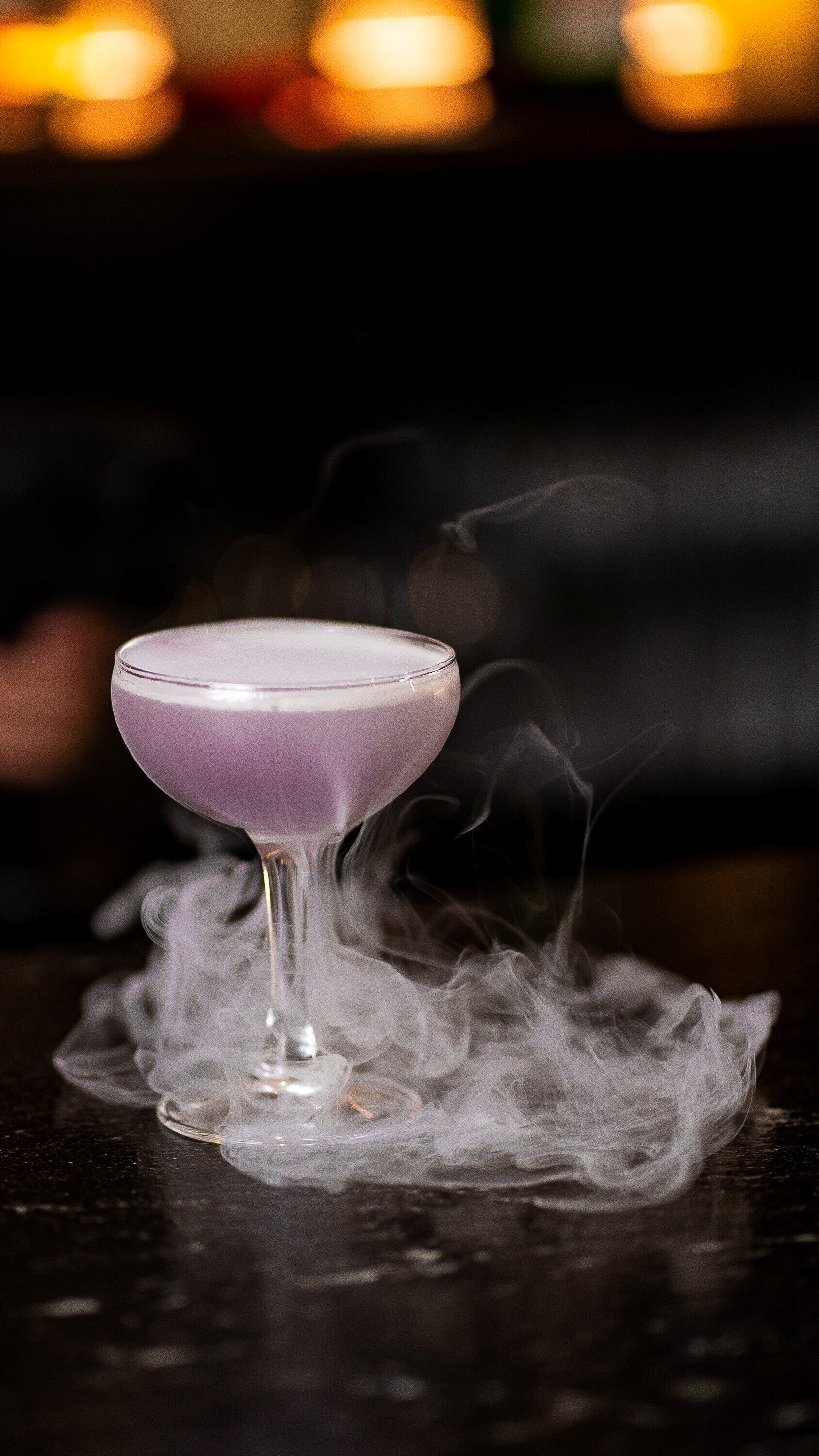 A purple cocktail with smoke rising from it, placed on a dark surface.