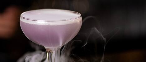 A purple cocktail with smoke rising from it, placed on a dark surface.