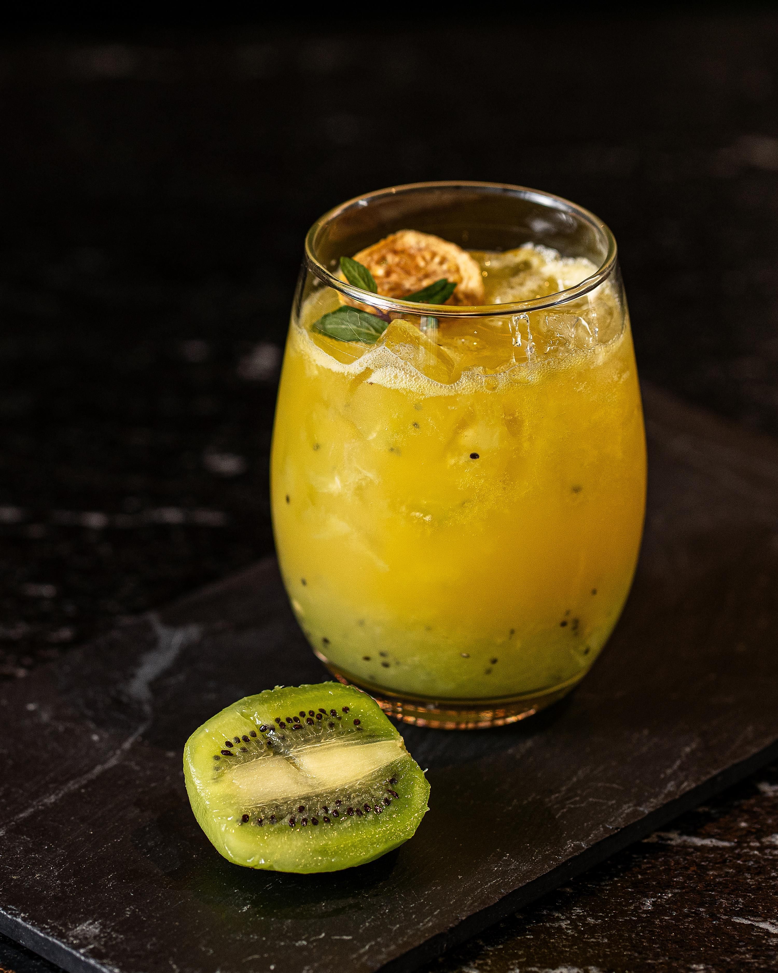 A glass of yellow beverage with ice cubes, garnished with a mint leaf, on a dark surface.
