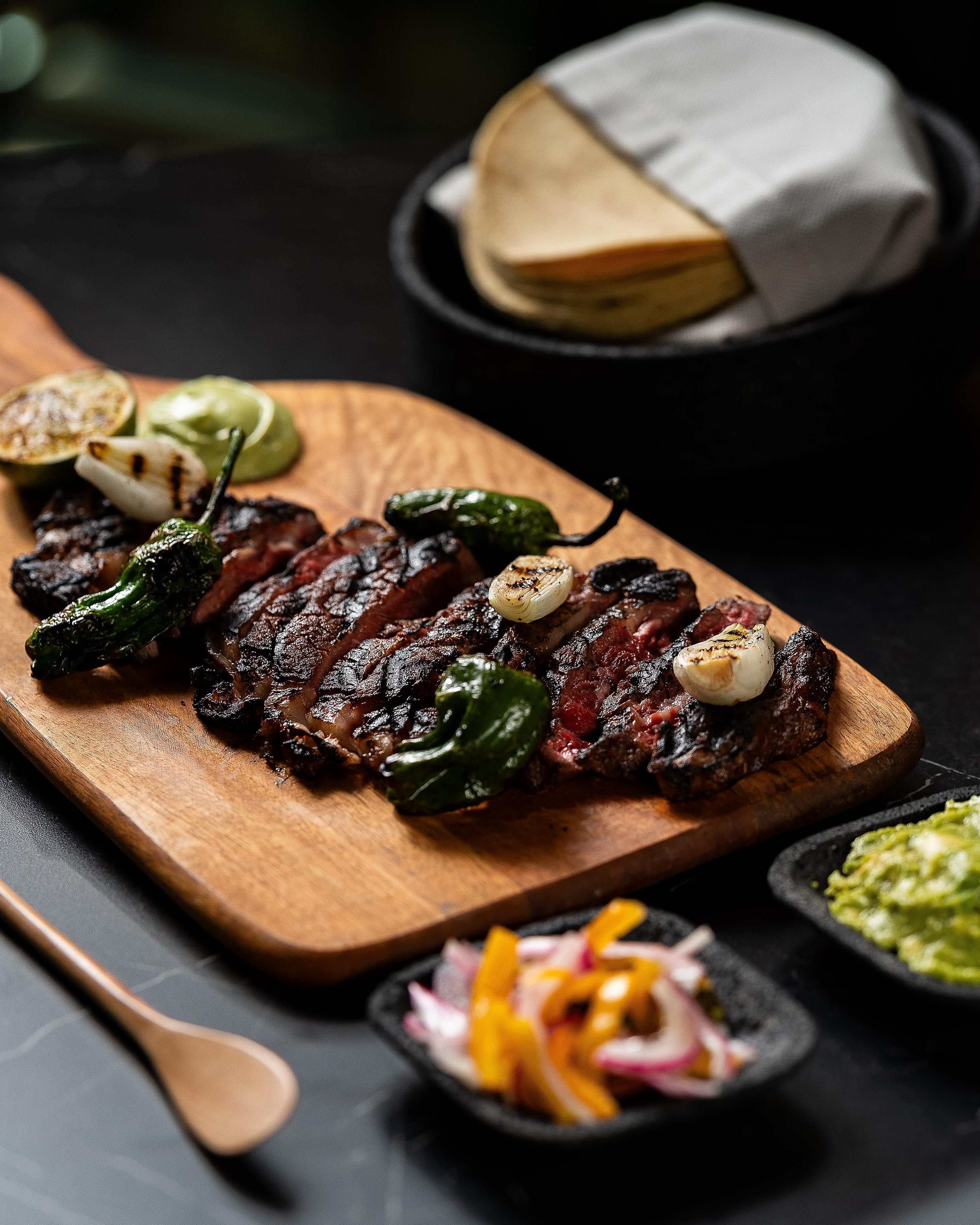 Una tabla de madera con carne a la parrilla, verduras y un recipiente pequeño con salsa.