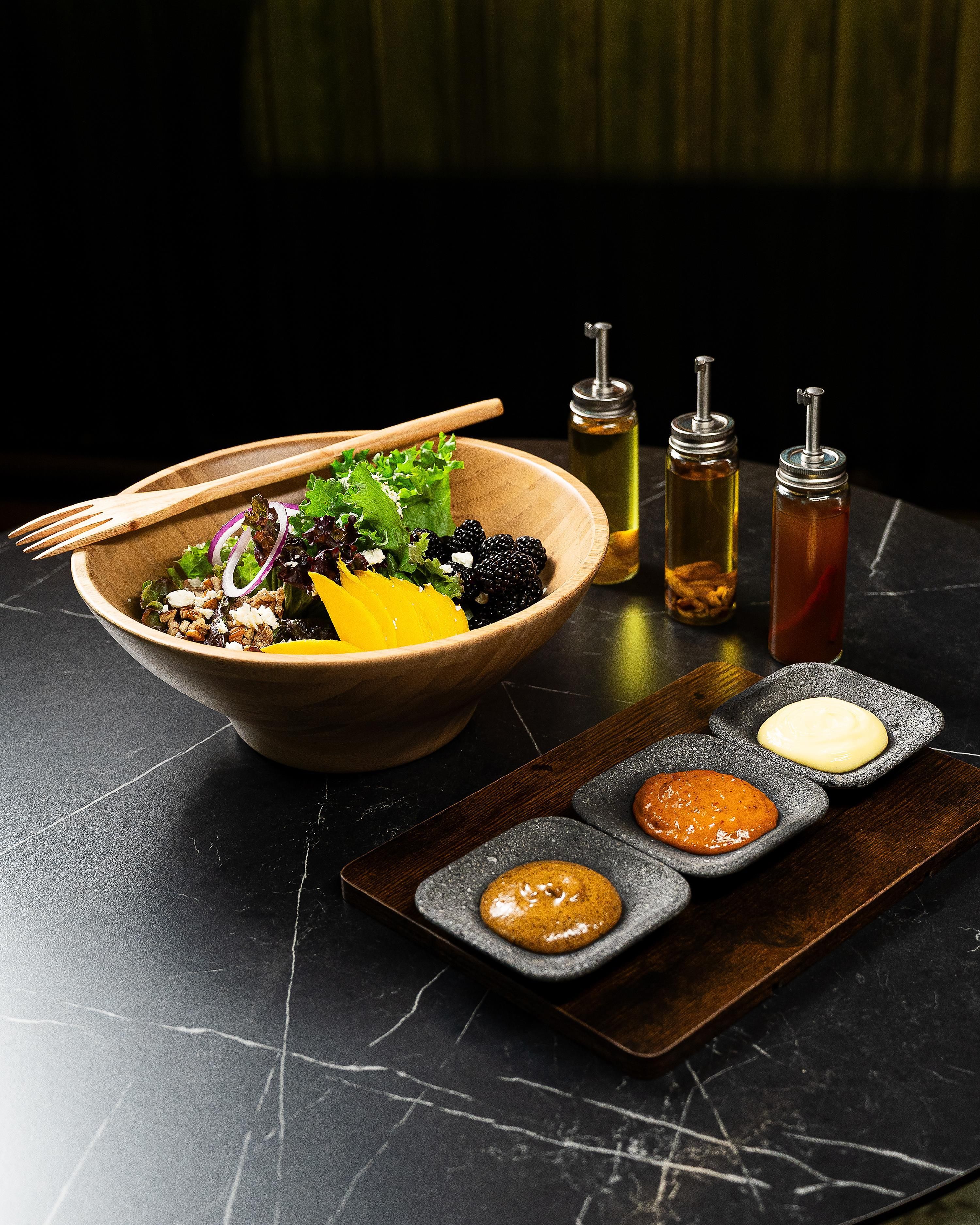 Un cuenco de madera con una ensalada, acompañado de tres recipientes pequeños de salsas sobre una superficie oscura.