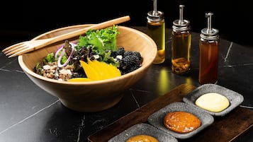 Un cuenco de madera con una ensalada, acompañado de tres recipientes pequeños de salsas sobre una superficie oscura.