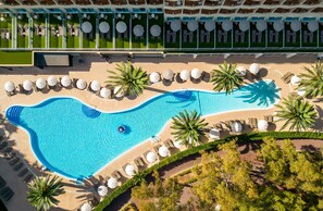 Ovenfra ses et resort med en stor swimmingpool formet som en fisk, omgivet af palmer og liggestole.