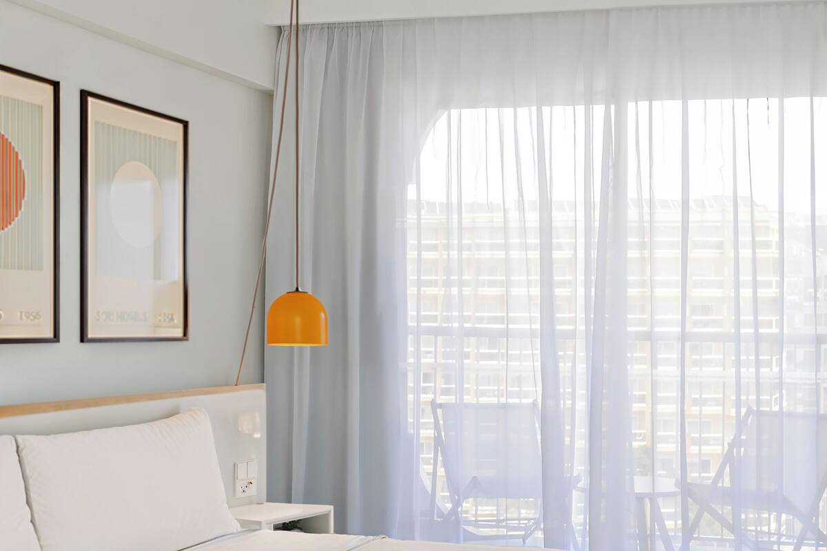 Habitación de hotel con una cama grande, una mesita de noche, una lámpara y vistas de la ciudad a través de cortinas translúcidas.